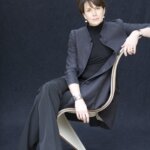 Woman, Davina Mallinckrodt, with short hair, sitting in a white chair in grey suit and black top and heels.