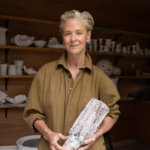White woman with grey hair, Deborah Brett, smiling holding a ceramic vase and wearing a green jumpsuit