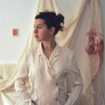 A woman, Lucrezia Abatzoglu, in a white shirt looking to the left with white drapery hung against a white wall behind her.