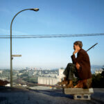 Man sitting on top of a block on a road which overlooks a city horizon with lamppost.