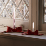 Two red bow ceramic candle holders with pink and white candles, sitting on a windowsill in front of a window.