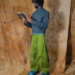 Photograph of a man, YAKU, in baggy green trousers and blue jacket and denim shoes. He is stood in front a beige background with doodles and reading a leather notebook.