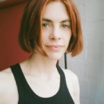 Pale white woman, Rebecca Ackroyd, with short red hair smiling in front of a red door wearing a black tank top