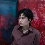 Asian woman, Amy Hui Li, in a maroon shirt, looking to the side. The woman is sat in front of two paintings which are red and blue.