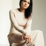 Woman, Saskia Fernando, with brown hair. She is wearing a beige dress and sitting down with a beige background.