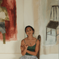 Woman, Layla Andrews, in a green tank top, sitting in a chair with two paintings behind