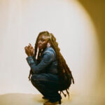 Black woman, Heather Agyepong, wearing a blue denim jumpsuit and red lipstick. She is crouching down with her hands held with a spotlight on her.
