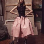 Woman, Florence Houston, wearing a baby pink skirt and a black waistcoat. Her hands are on her hips and she is sitting down in a studio.