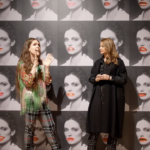 Two woman, talking in front a wall with multiple versions of black and white photo of a woman with large red lips.