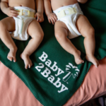 Two babies legs wearing diapers, lying on a green blanket on a pink duvet. On the green blanket it says Baby 2 Baby