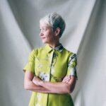 Woman, Maria Balshaw, with short grey hair, looking to the side. The woman is standing in front of a white backdrop, wearing a lime green dress with flowers and red lipstick. Her arms are crossed over.