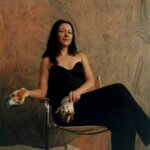 A woman with brown hair wearing a black jumpsuit, holding gloves covered in paint sat in a chair