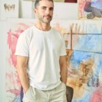 A man wearing a white t-shirt and beige trousers, standing in front of paintings
