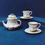 Two white teacups with detailing a white teapot with the word written white on in.