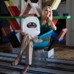 A woman sat in a chair with blonde hair in front of a colourful bookshelf