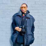 Black woman wearing a black jacket with orange sunglasses holding a camera