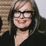 A woman smiling for a photograph with grey hair and black glasses