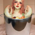 A woman sitting in a silver bucket with pink hair and lobster hands