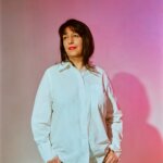 A woman wearing a white shirt standing in front of a pink background.