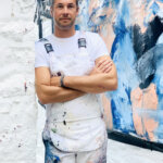 Artist Wesley Eberle leaning against a white brick wall with an abstract blue painting in the background