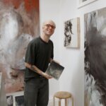A man in glasses posing in an artist studio with an artwork in his hands.