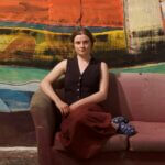 A female artist sitting on a sofa in front of painted artwork.