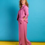 A red-head lady in a pink suit stands in front of a turquoise background on a yellow floor.